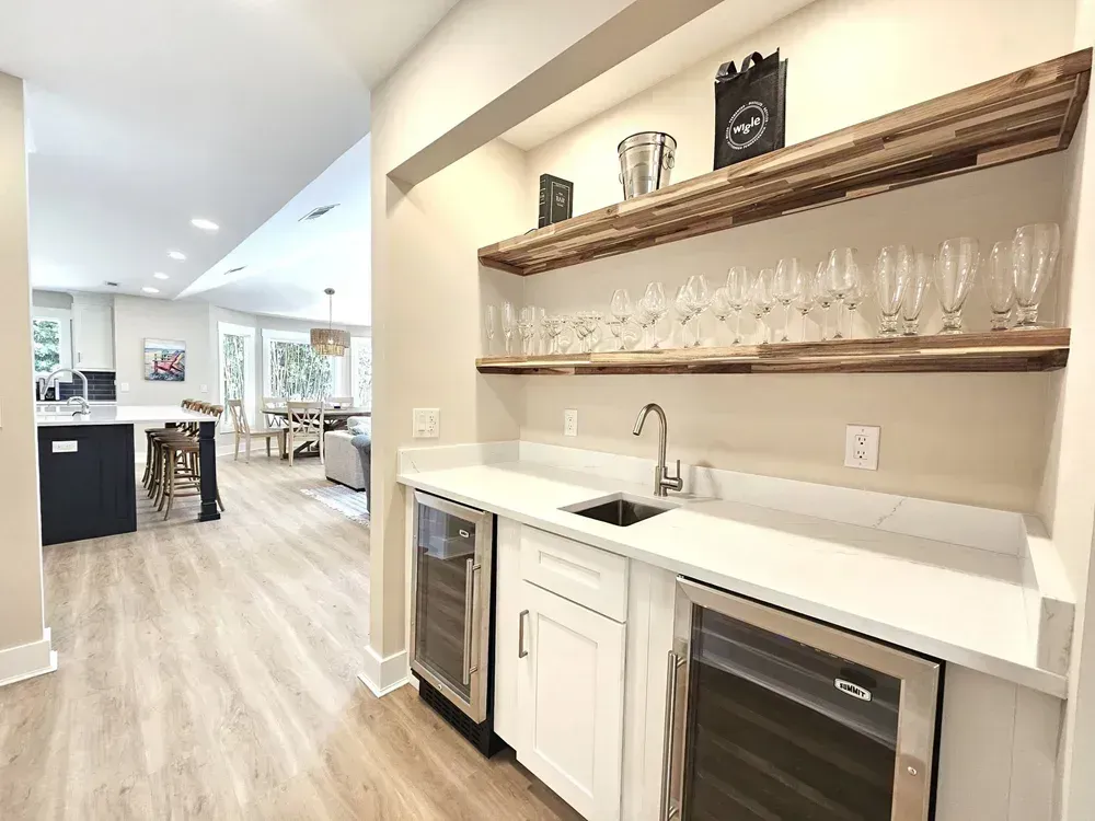 A built-in wet bar with white quartz countertops, a stainless sink, two glass-front beverage coolers, and floating wood shelves lined with stemware, opening onto a kitchen and living area with light wood flooring.