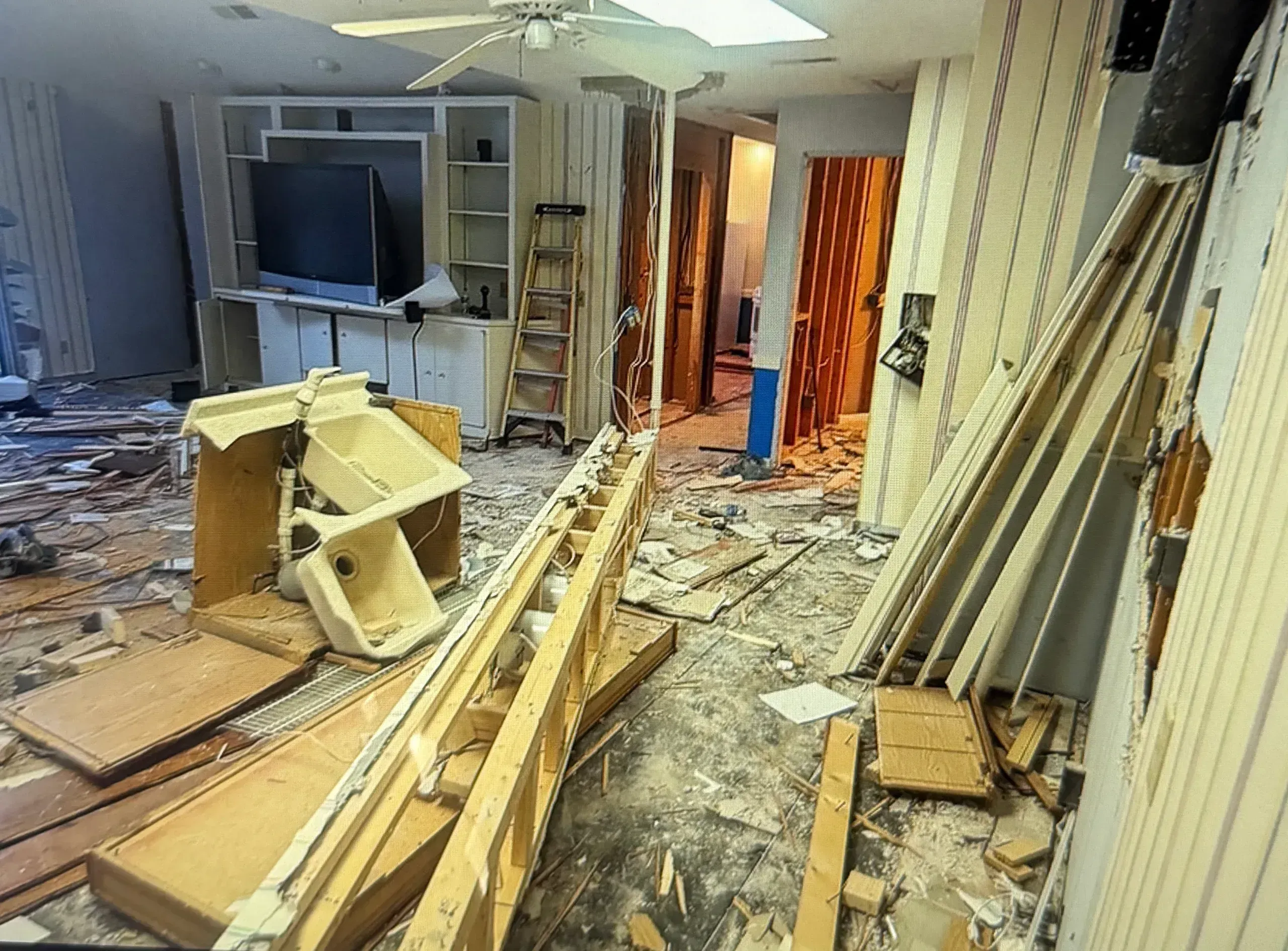 An interior room mid-demolition shows exposed wall studs, a detached wooden vanity with sink, scattered debris, and a stepladder near a built-in entertainment center.
