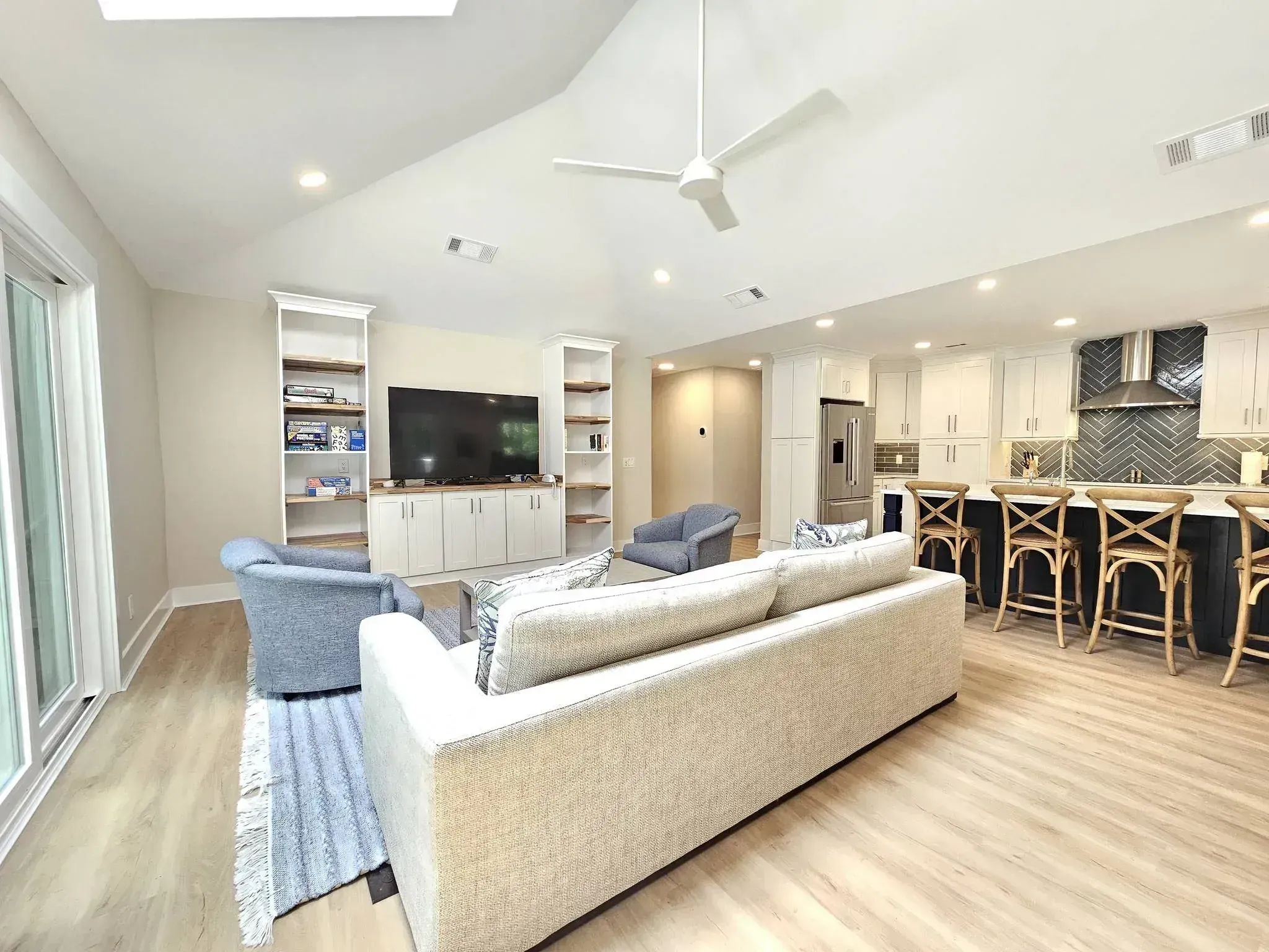 An open-concept living room with a vaulted ceiling, ceiling fan, and skylight flows into a kitchen with white cabinetry, a herringbone tile backsplash, and a dark island lined with woven barstools.
